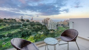 A balcony with wicker chairs and a glass table overlooking a cityscape with buildings and greenery. - Valley Sea Views With Furnished BBQ Balcony (St. Julian's)