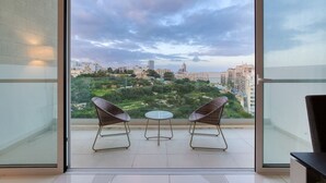 A balcony with two wicker chairs and a small table, overlooking a cityscape with buildings and greenery. - Valley Sea Views With Furnished BBQ Balcony (St. Julian's)