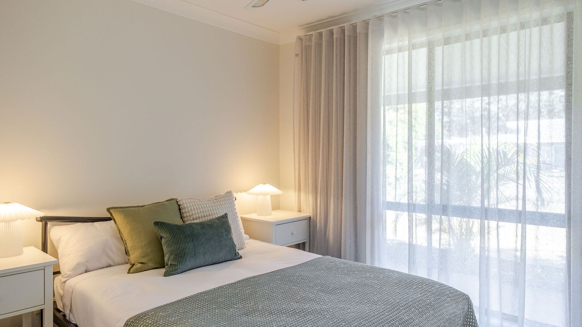 A hotel room with a bed, two bedside lamps, a window with sheer curtains, and a view of trees outside.