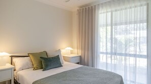 Une chambre d’hôtel avec un lit, deux lampes de chevet, une fenêtre avec des rideaux légers et une vue sur les arbres à l’extérieur.