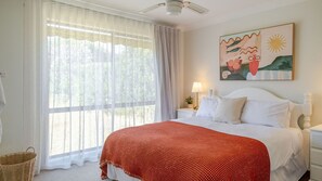 A bedroom with a bed, a nightstand, a lamp, a painting, and a window with curtains. - Narrawallee Nirvana - Beach House (Narrawallee)