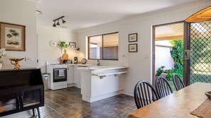 A compact kitchen with a central island, a dining area with a wooden table, and a view of a garden through a glass door. - Narrawallee Nirvana - Beach House (Narrawallee)