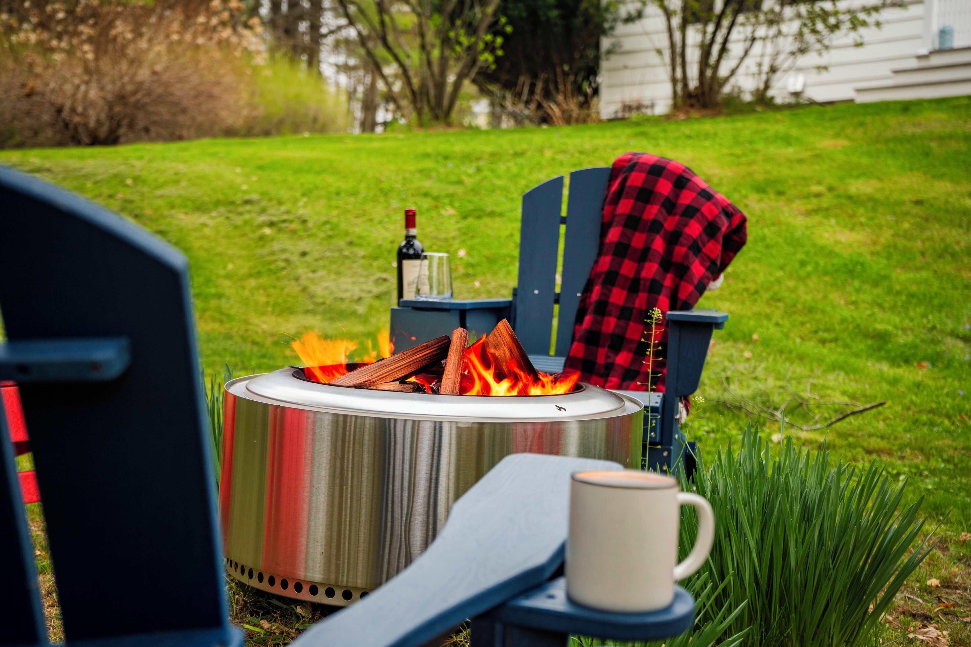 A portable fire pit with logs, a bottle, and a glass on a stand, placed on a lawn with a chair and a mug nearby.
