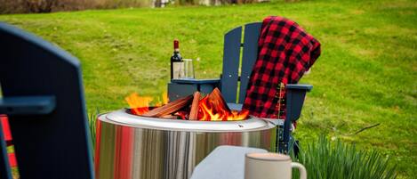 A portable fire pit with logs, a bottle, and a glass on a stand, placed on a lawn with a chair and a mug nearby.