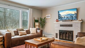 A cozy living room with a fireplace, a sofa, a coffee table, and a large window.