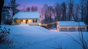 A snow-covered house with a front porch and a garage, surrounded by trees and a snowy landscape.