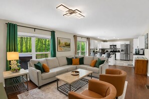 Ein modernes Wohnzimmer mit Sofa, Couchtisch und Sesseln, eine Küche mit Edelstahlgeräten und großen Fenstern mit grünen Vorhängen.