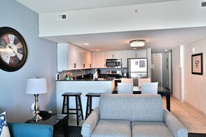 A modern living room with a grey sofa, a dining table with chairs, a kitchen area with white cabinets and appliances, and a wall clock. - Discover comfort and coastal charm in this 3-bedroom Myrtle Beach condo (Myrtle Beach)