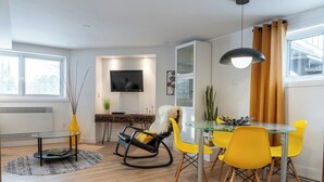 Een moderne woonkamer met een glazen eettafel, gele stoelen, een zwarte schommelstoel, een aan de muur bevestigde televisie en een groot raam met gordijnen.