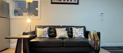 A modern living room with a black leather sofa, patterned cushions, a small table with a lamp, and a framed artwork on the wall.