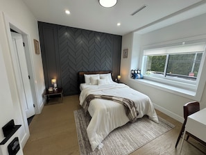 Une chambre avec un mur orné d’un motif en chevrons, un grand lit avec une literie blanche, une table de chevet avec une lampe, une fenêtre donnant sur un paysage agréable et un plancher en bois.