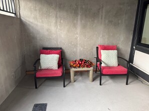 Zwei Stühle mit roten Kissen und weißen Kissen, ein kleiner Tisch mit einer Blumenkomposition und ein Betonboden.