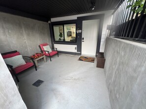 A covered outdoor seating area with two chairs and a bench, a door, and a concrete wall. - Luxe 2BD Suite/Sweet Spot in South Surrey (Surrey)