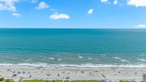 A beach with a clear sky, calm sea, and a sandy shore with umbrellas and people. - Ocean Villas 502 (Myrtle Beach)