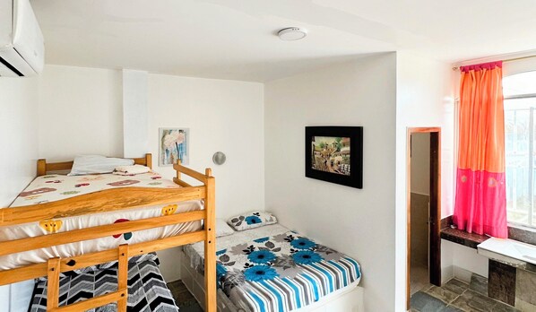 A bedroom with a bunk bed, a single bed, a small bedside table, a window with curtains, and a picture on the wall.
