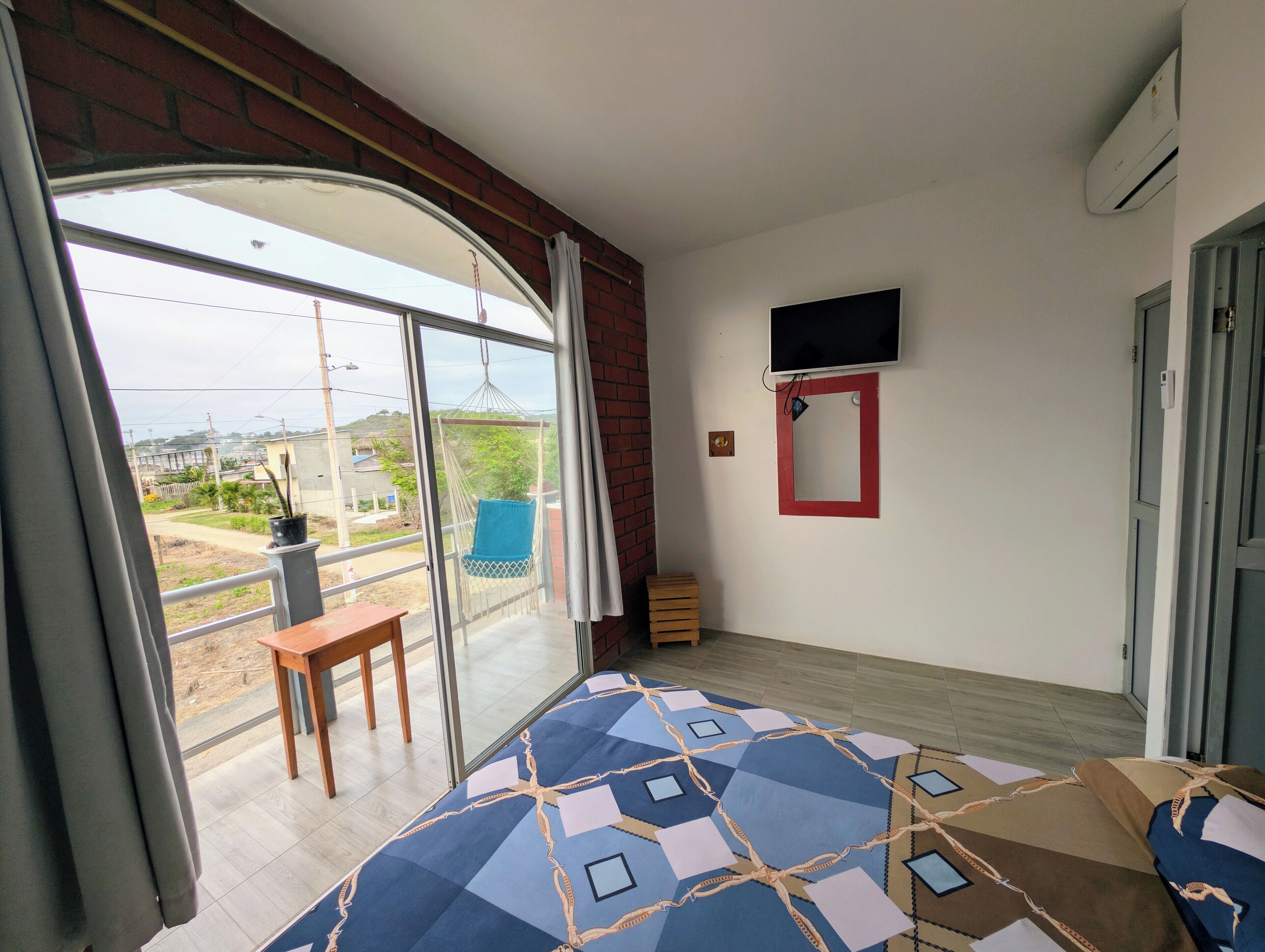 Una habitación con un gran ventanal corredero que da a un balcón, una cama con colcha de estampado geométrico, una pequeña mesa de madera, un televisor montado en la pared y un espejo con marco rojo.