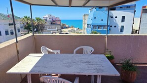 Un balcon avec une table et des chaises, donnant sur une zone côtière avec des bâtiments et la mer.