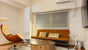 Uma sala de estar moderna com uma mesa de centro de madeira curvada, um sofá laranja com capitonê e duas almofadas, uma cadeira de madeira e uma pequena mesa lateral branca.