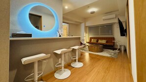 Um banheiro moderno com um espelho circular, três banquetas brancas, uma penteadeira de madeira e um espaço de estar com sofá e mesa de centro.