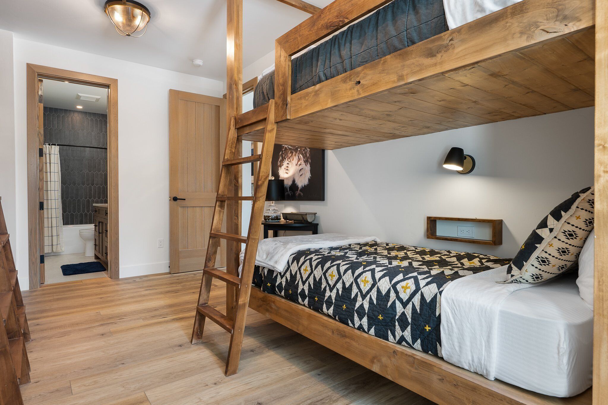 A bunk bed room with a wooden ladder, a desk with a lamp, and a bathroom visible through an open door.