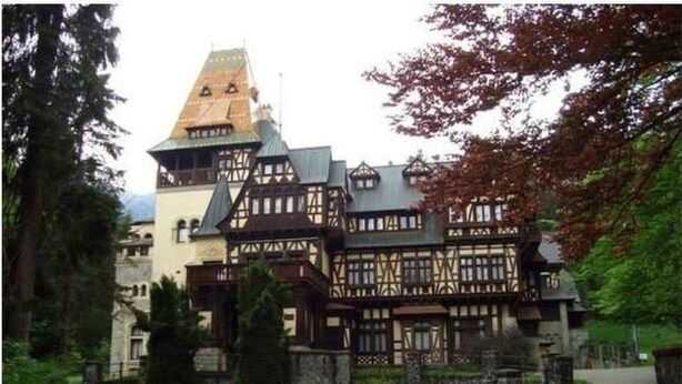 Un edificio tradicional con entramado de madera y una torre destacada, rodeado de abundante vegetación y un camino de grava.