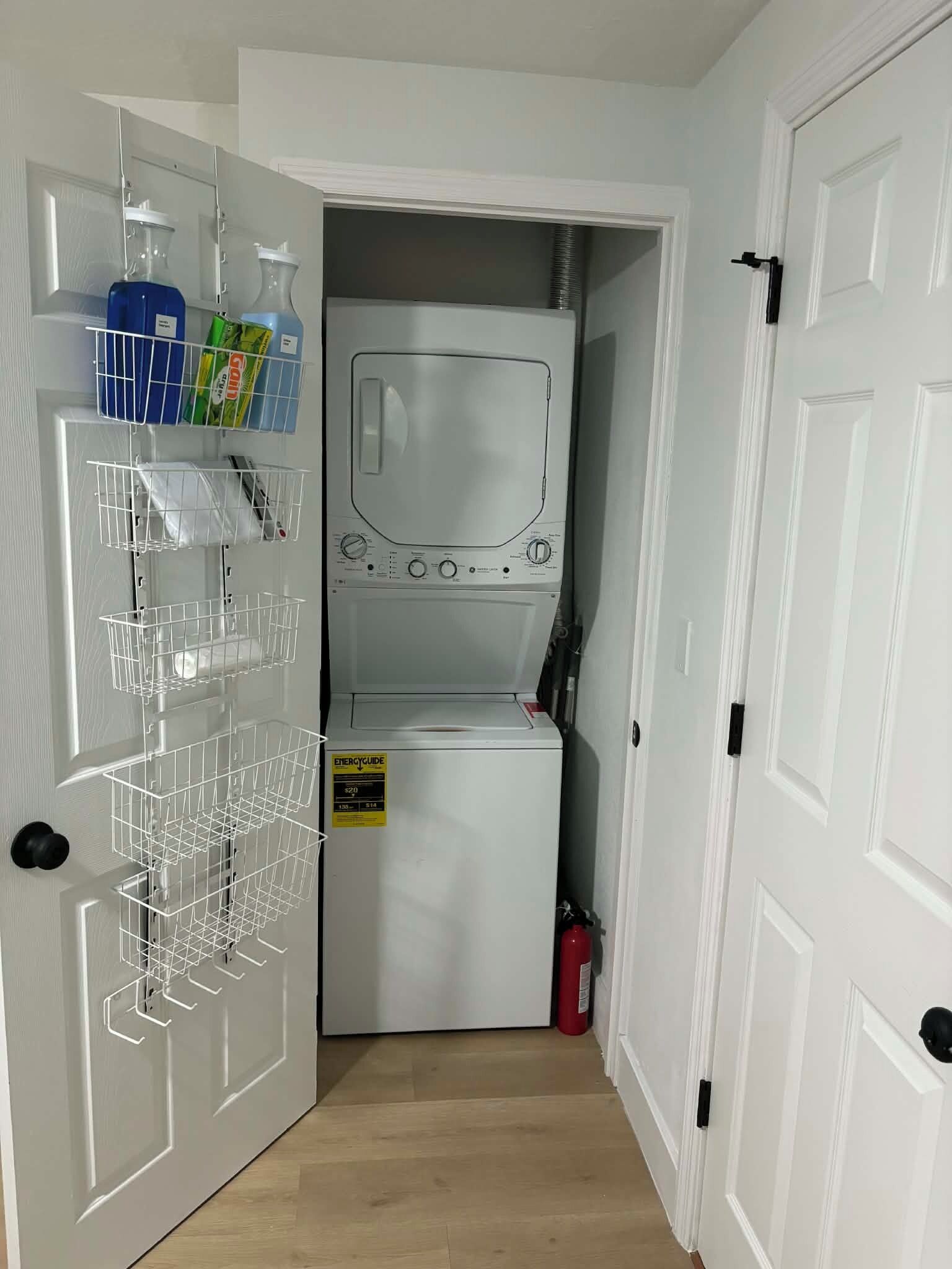 A laundry room with a white washing machine, a dryer, and a metal rack with hanging clothes. There are storage shelves above the machine and a fire extinguisher to the right.