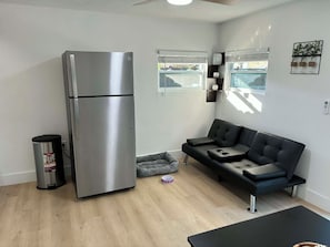 Ein kompakter Wohnraum mit einem Edelstahl-Kühlschrank, einem schwarzen Sofa, einem grauen Tierbett und einer kleinen Mülltonne.