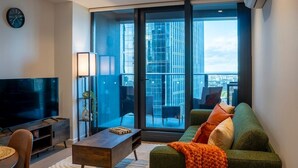 A modern living room with a large flat-screen TV, a sofa with colorful cushions, a coffee table, and a view of a cityscape through large glass doors. - Luxe 2BR Escape | City View, Pool & Gym (Docklands)