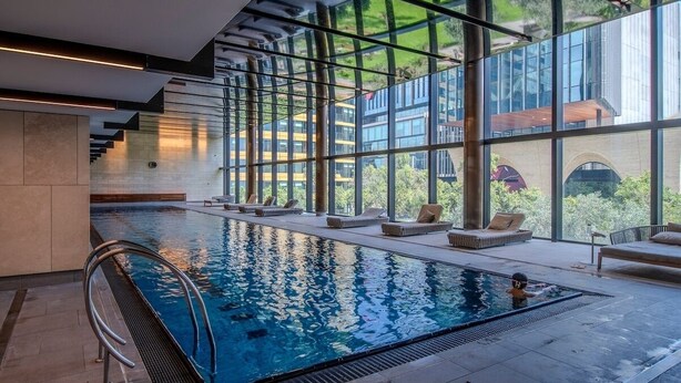 A modern indoor swimming pool with clear glass walls, lounge chairs, and a view of the surrounding buildings. - Luxe 2BR Escape | City View, Pool & Gym (Docklands)