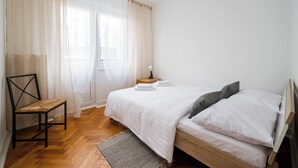 A bedroom with a wooden bed, white bedding, a chair, a bedside table with a lamp, and sheer curtains. - Twarda Downtown Apartment (Warszawa)