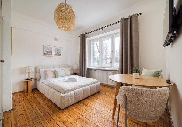 A bedroom with a wooden floor, a bed with a white comforter, a round wooden table with a chair, a window with curtains, and a wall-mounted TV. - Saska Kępa Lavish Studio (Warszawa)