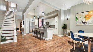 Una cocina amplia con una isla central, un comedor con sillas azules y una escalera que lleva a otro nivel.