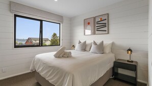 Une chambre avec une grande fenêtre, un lit recouvert de draps blancs, des oreillers à rayures et une table de chevet avec une lampe.