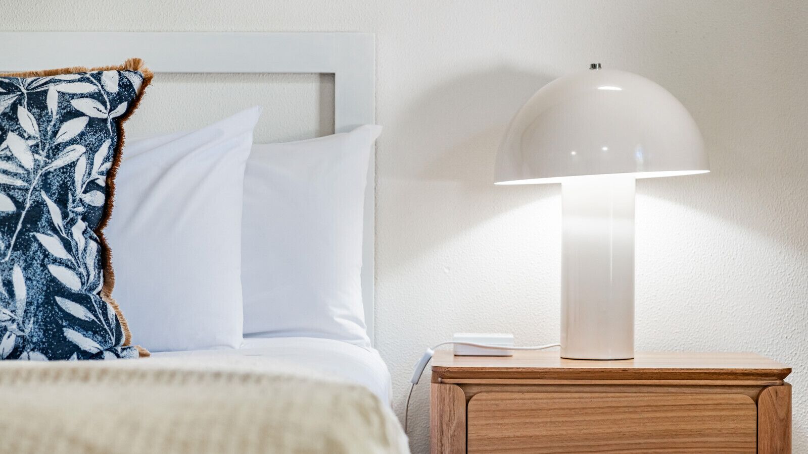 Una cama bien tendida con almohadillas blancas y azules estampadas, una manta blanca y una mesita de noche de madera con una lámpara blanca.