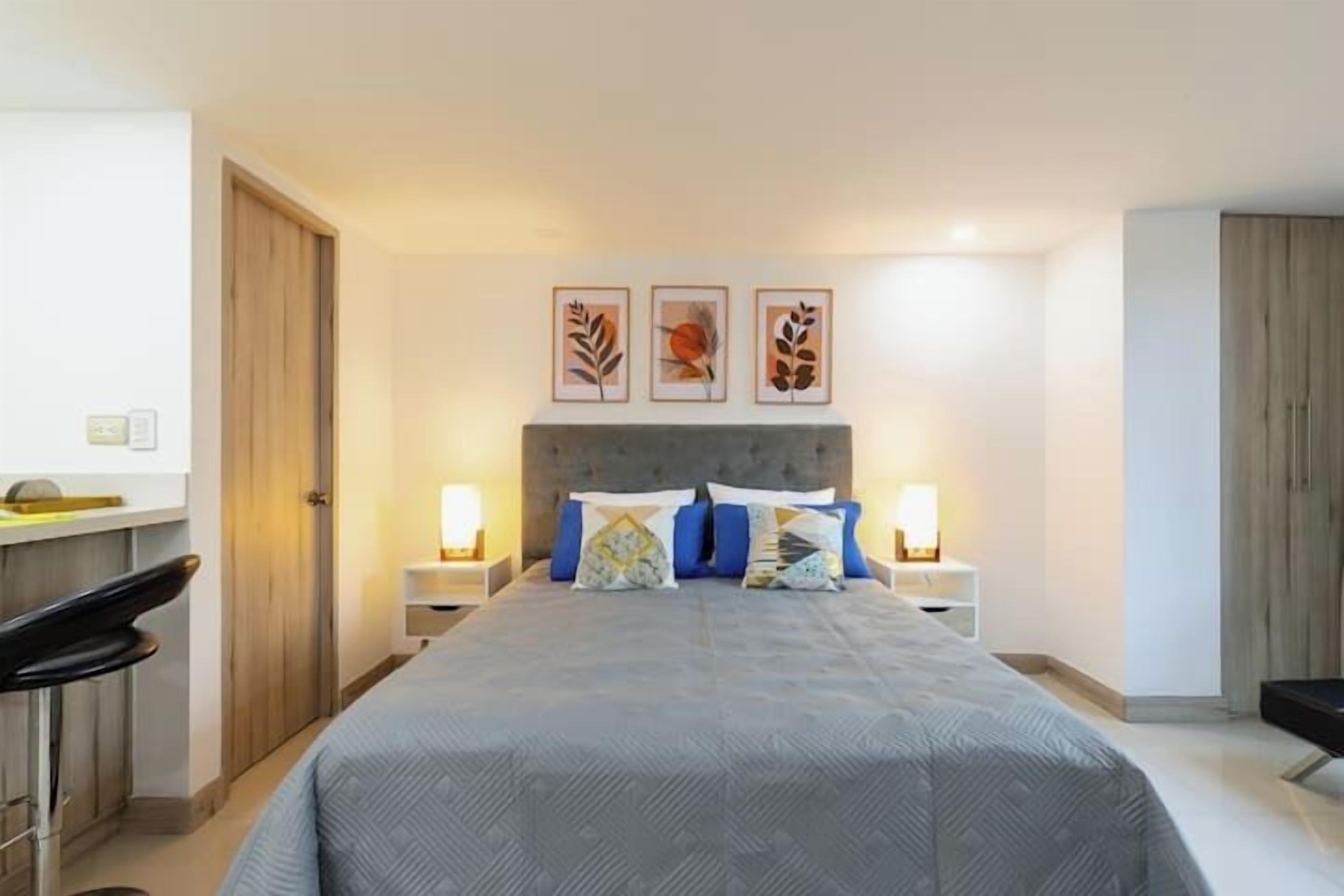 A modern bedroom with a large bed, two bedside tables with lamps, a desk with a chair, and three framed artworks on the wall.