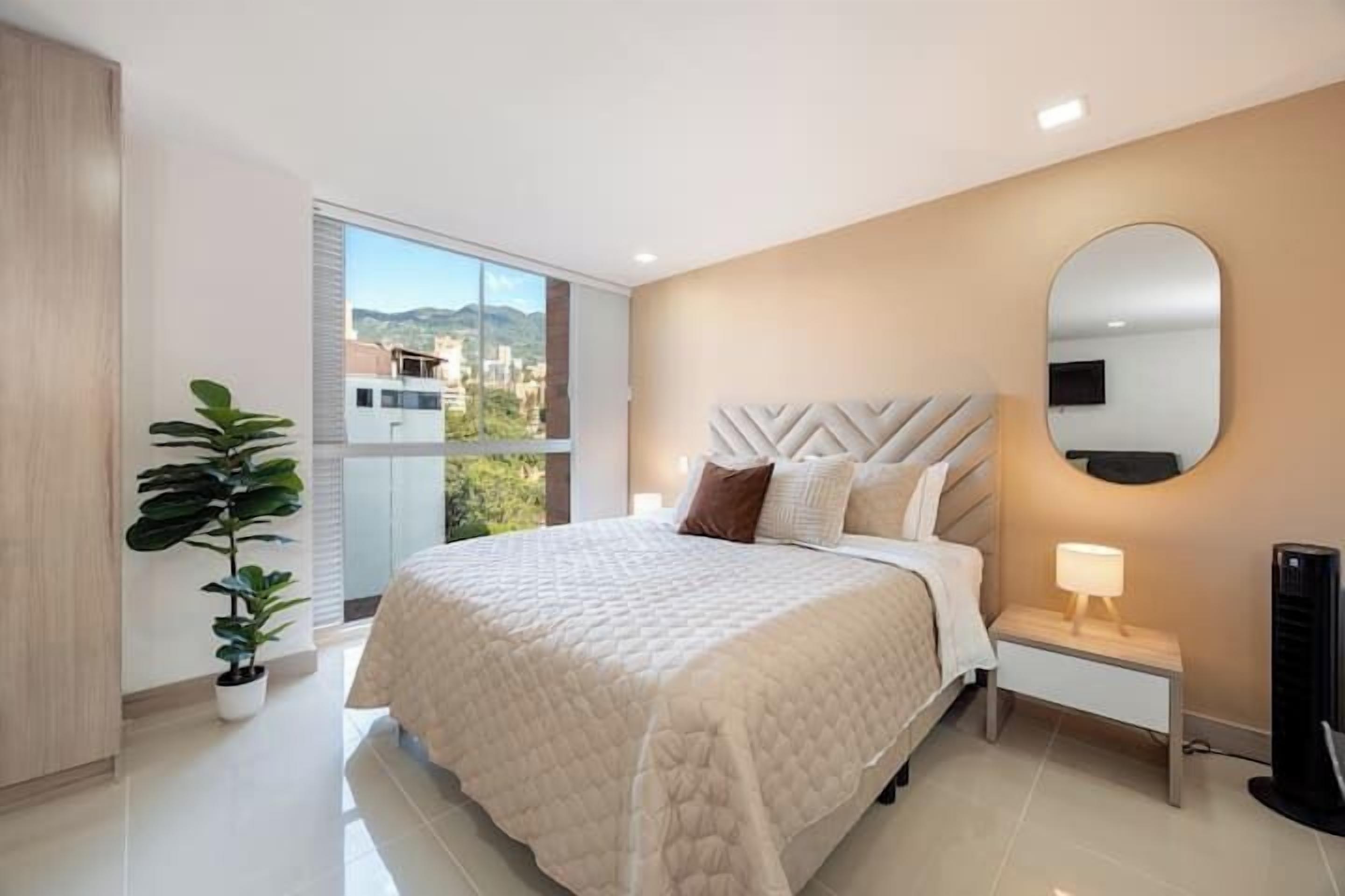 A modern bedroom with a large bed, a bedside table with a lamp, a potted plant, and a large mirror on the wall.
