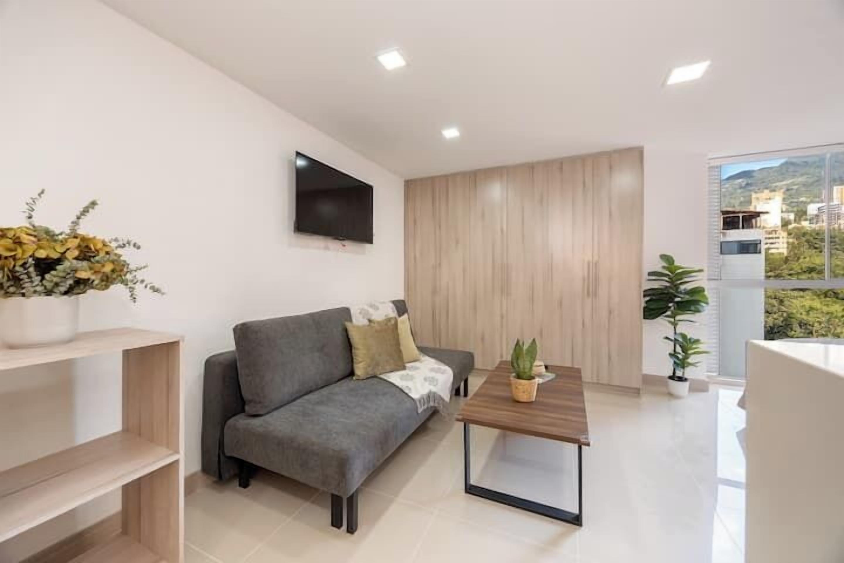 Ein modernes Wohnzimmer mit grauem Sofa, einem Holztisch, einem flachbildfernseher an der Wand und einem großen Fenster mit Blick auf Gebäude und Grünflächen.