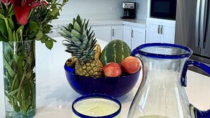 Et køkken med hvide skabe, en rustfrit stål køleskab, en skål med frugt, en kande og et glas cocktail.
