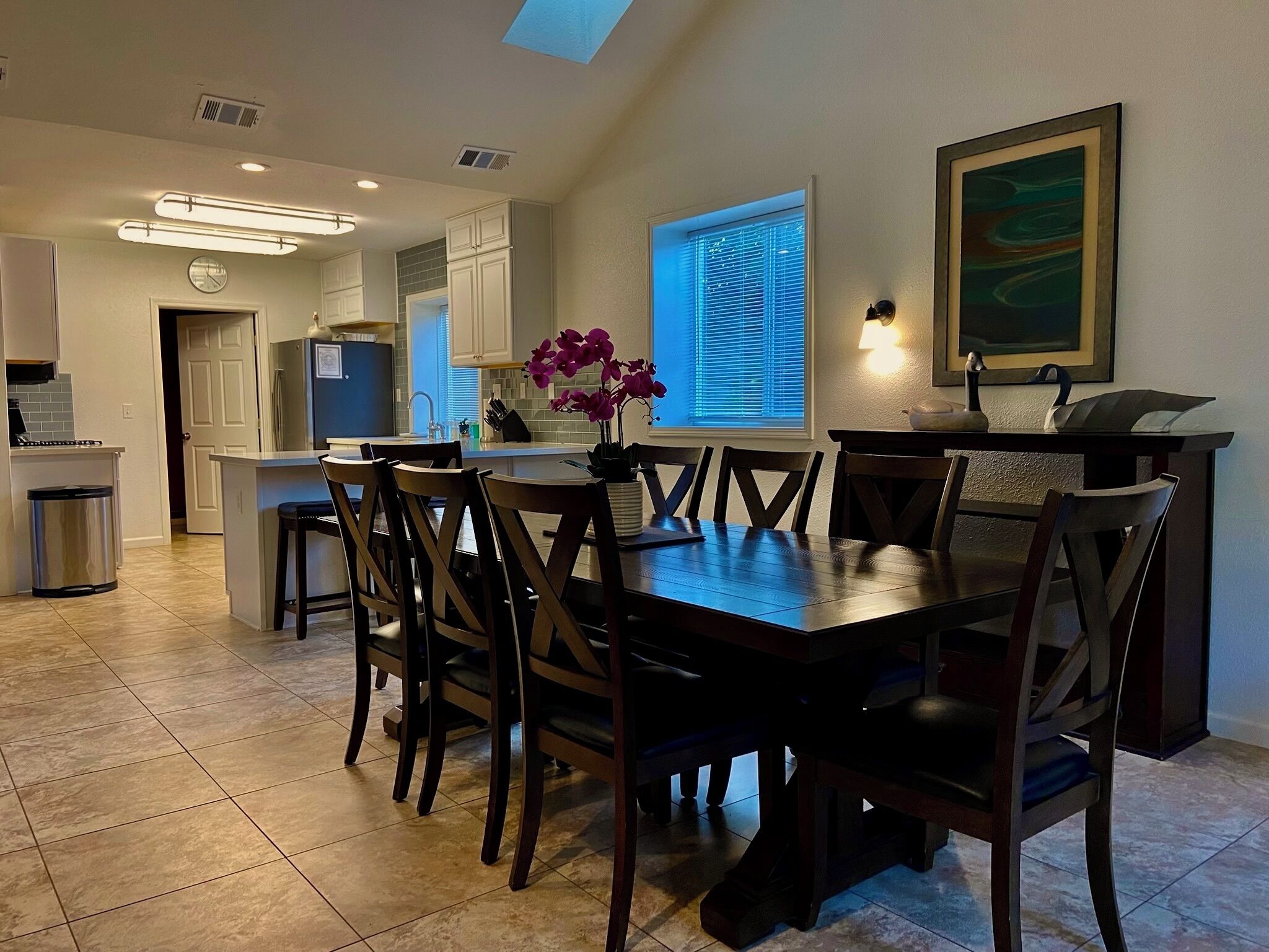 Un comedor con una mesa y sillas de madera oscura, una cocina al fondo, un bote de basura y una ventana con persianas.