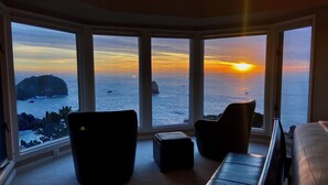 Een kamer met een grote erker die uitkijkt op de oceaan bij zonsondergang, met een bank, stoelen en een klein voetenbankje.