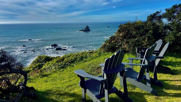 A view of the ocean with waves crashing onto rocks, two Adirondack chairs facing the water, and lush greenery in the foreground. - Trinidad Coastal Retreat – Open Living, Panoramic Ocean Views & Stylish Comfort (Trinidad)