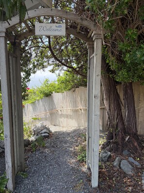 A wooden archway with a 'Welcome' sign, surrounded by greenery and a gravel path.