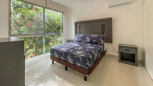 Ein Schlafzimmer mit Bett, Nachttisch, Deckenventilator und einem Fenster mit Blick auf Grünflächen.