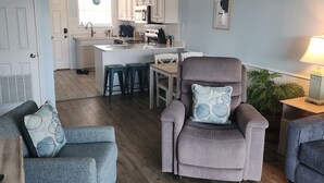 A living room with a grey sofa, a grey armchair, a ceiling fan, and a kitchen area in the background. - The Shore Thing - Gulf Shores (Gulf Shores)