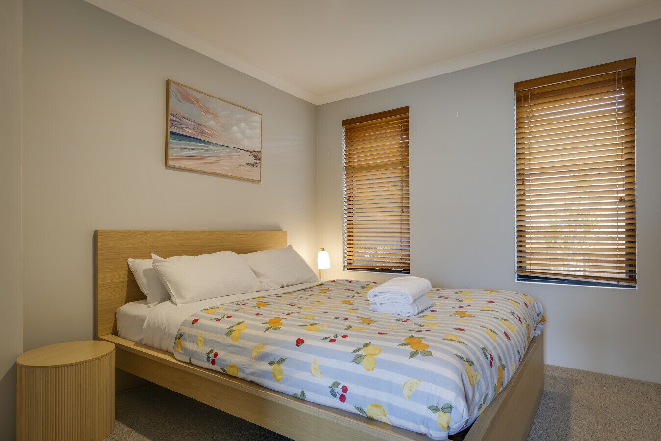 Un dormitorio con una cama, una lámpara de noche, una mesita redonda y dos ventanas con persianas.
