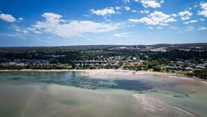 Ett flygbildsbild av ett kustnära område med klart vatten, en sandstrand och ett bostadsområde med hus och grönområden.