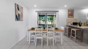 Un coin repas avec une table en bois et des chaises blanches, une cuisine équipée d’un four à micro-ondes et d’un évier, et une vue sur la verdure environnante.