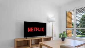 Un salon moderne avec un téléviseur à écran plat affichant le logo de Netflix, un meuble TV en bois, une lampe sur pied, une table à manger et une plante en pot.