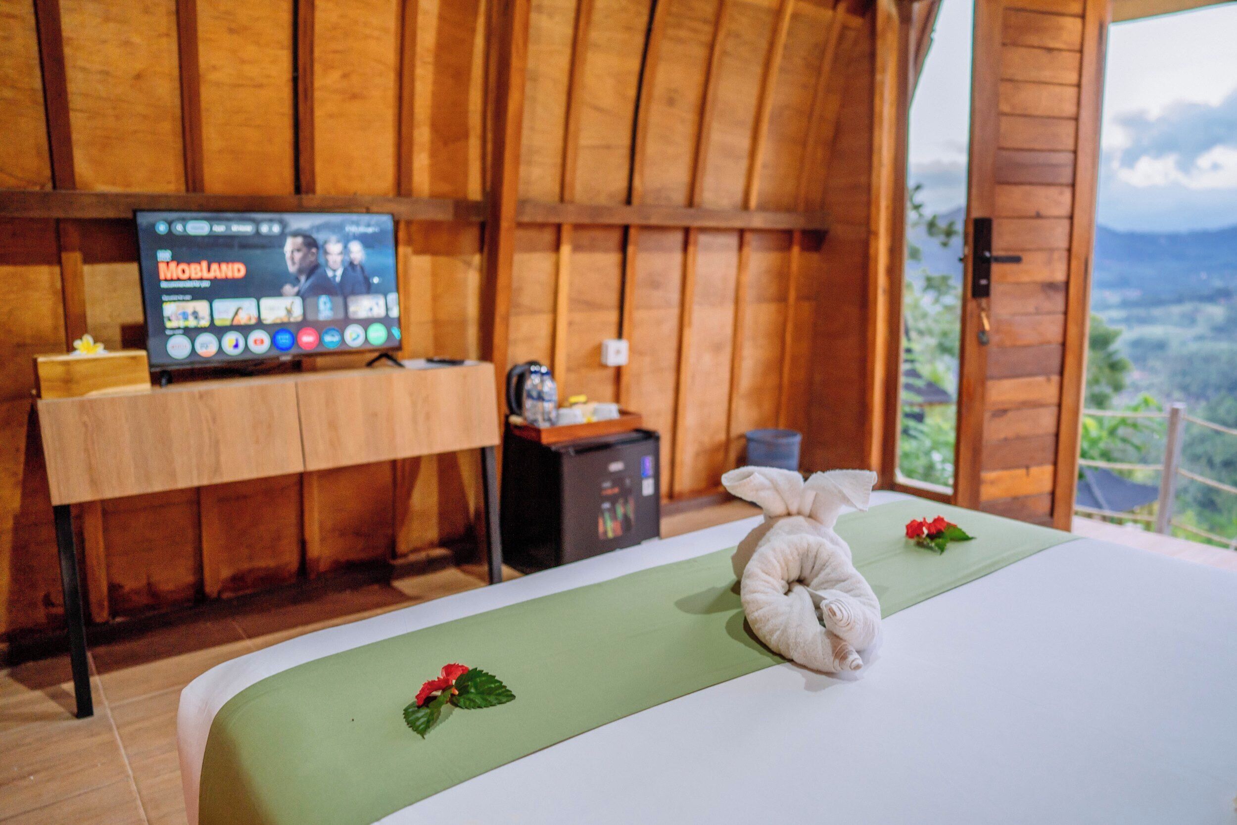 Een kamer met een bed, een televisie waarop 'MOBILAND' te zien is, een klein tafeltje met een lamp, en uitzicht op het omliggende landschap.