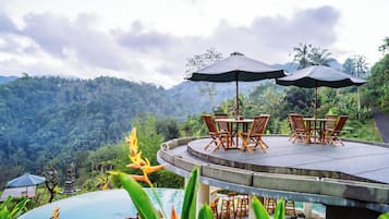 A terrace with a table and chairs, surrounded by lush greenery and a mountainous backdrop.